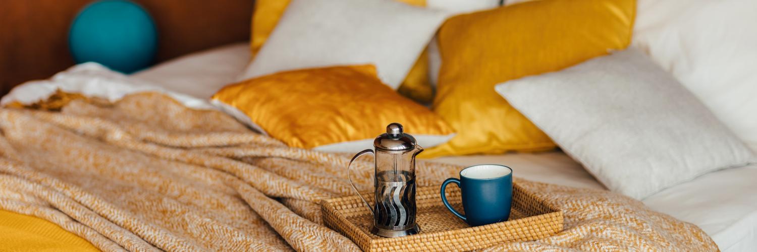 Tea with a cozy winter home background and a warm yellow knitted plaid and orange cushions on bed background. Bed with bedspread, white sheets and pillows with wooden tray and coffee cup. Sweet home. Breakfast in bed. Lifestyle concept.